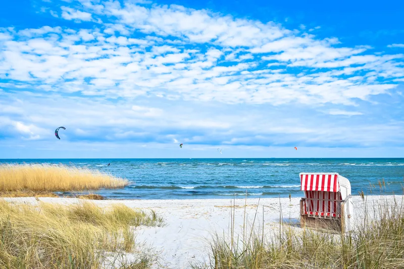 Rot-weiß gestreifter Strandkorb im Strandhafer am Ostseestrand, auf dem Wasser Kitesurfer unter weißen Wolken
